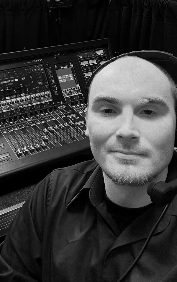 A man wearing headphones and a black shirt smiling in front of a professional audio mixing console.