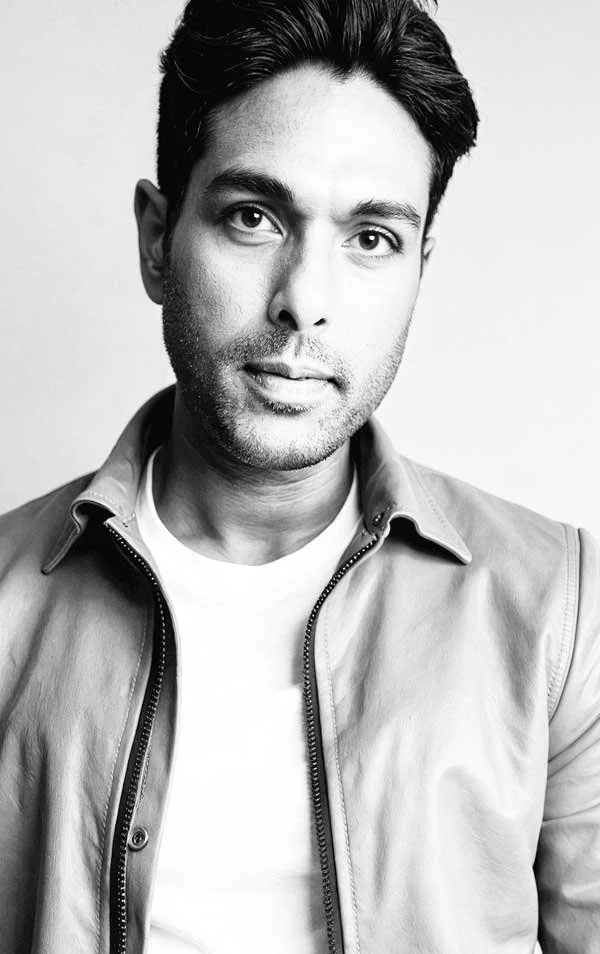 Black and white portrait of a man with dark hair, wearing a light-colored jacket and t-shirt, looking at the camera.