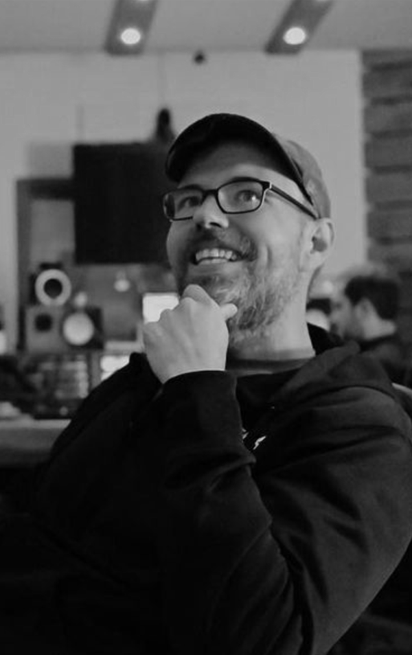 Man with glasses and a cap, smiling and resting his chin on his hand in a recording studio.