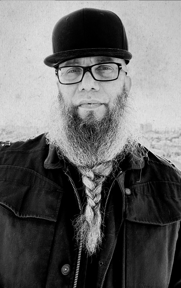 A man with a long, braided beard, glasses, and a black cap, wearing a dark jacket, standing outdoors against a textured wall.