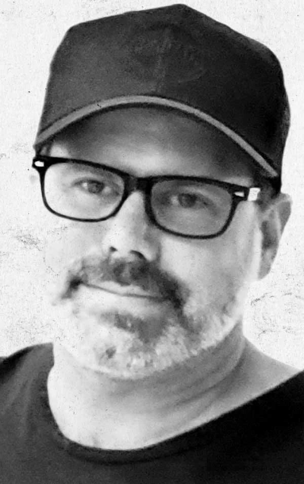 A man with glasses, a beard, and a mustache wearing a black cap and black shirt, smiling slightly in a black-and-white photo.