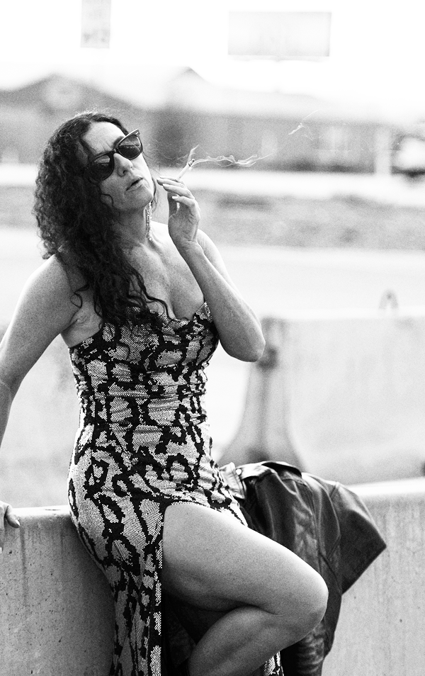 A woman in a snakeskin dress and sunglasses sitting on a barrier, smoking.