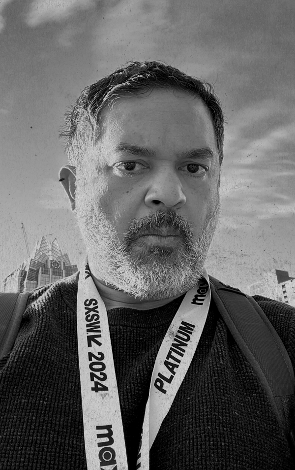 A man with a beard takes a selfie outdoors, wearing a lanyard with event details, and there are buildings and a cloudy sky in the background.