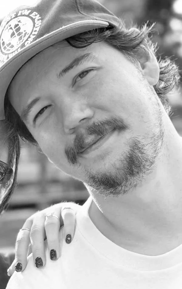 A young man with a beard and mustache wearing a baseball cap, smiling slightly, with a woman's hand resting on his shoulder.