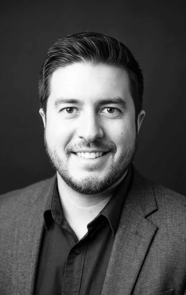 Black and white portrait of a young man with short, neatly combed hair, smiling, wearing a suit jacket and shirt against a dark background.