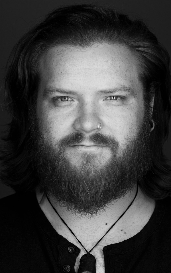 A black-and-white portrait of a man with long hair, a thick beard, and mustache, wearing a nose ring and a necklace.