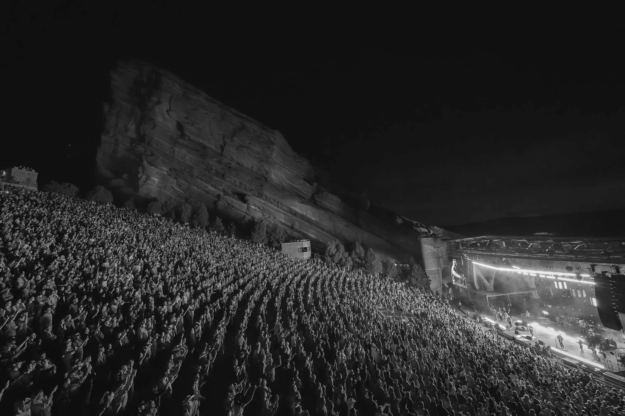 Black and white photo of a large outdoor concert at night with a full audience, stage lighting, and a rocky hillside in the background.