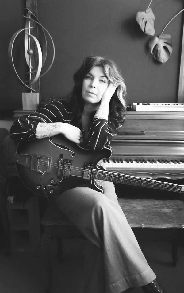 A woman sitting on a chair with an electric guitar, next to a piano, in a room with modern wall decorations.