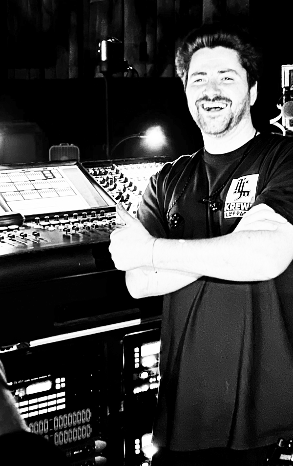 A man standing in a recording studio, smiling, with arms crossed, wearing a black T-shirt with a patch, standing next to audio mixing equipment.