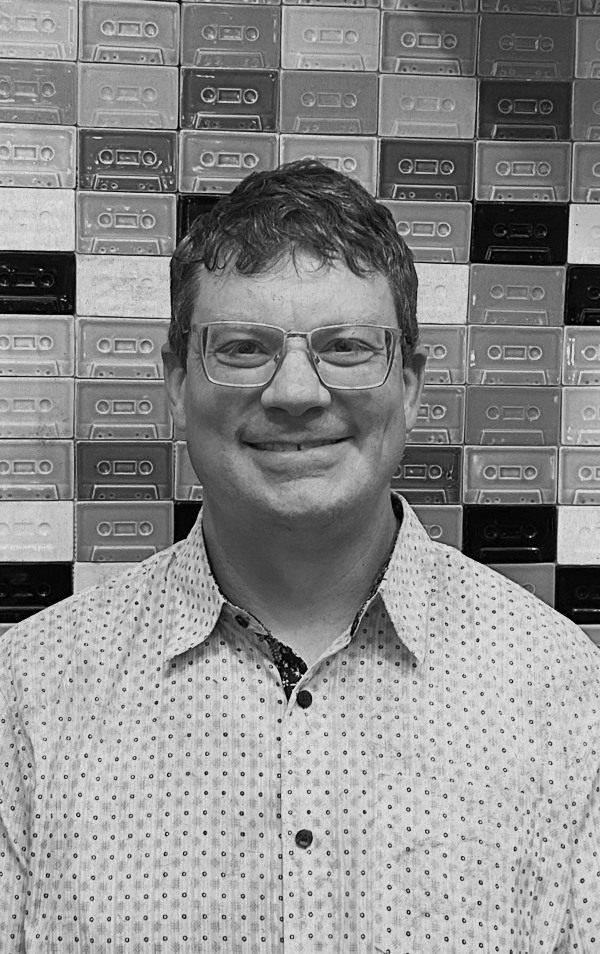 A man wearing glasses and a patterned button-up shirt is smiling in front of a wall of cassette tapes.