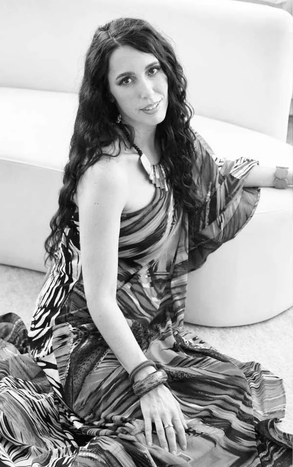 A woman with long wavy hair, wearing a patterned maxi dress, sitting on the floor beside a curved sofa, smiling at the camera.
