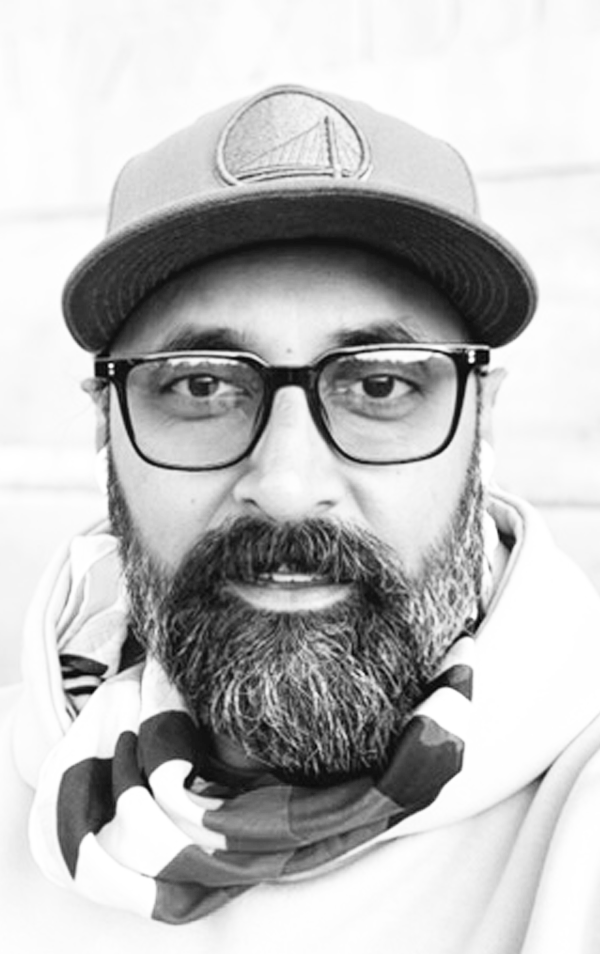 Close-up of a bearded man wearing glasses, a baseball cap with a logo, and a patterned scarf around his neck in black and white.