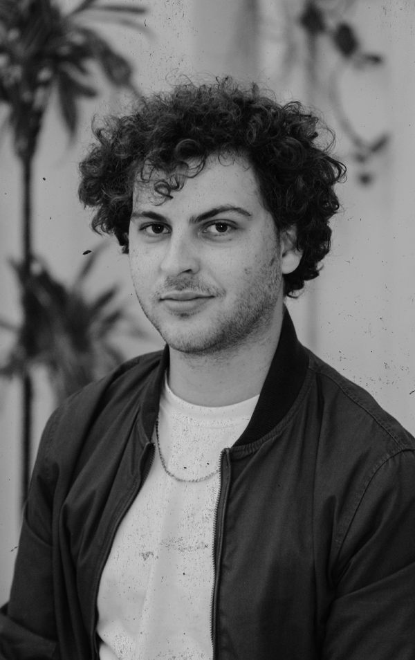 Black and white portrait of a young man with curly hair, wearing a bomber jacket over a white T-shirt, seated indoors with plants in the background.