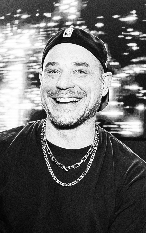 A smiling man wearing a beanie with a logo, layered necklaces, and a dark shirt, with a blurred cityscape background in black and white.