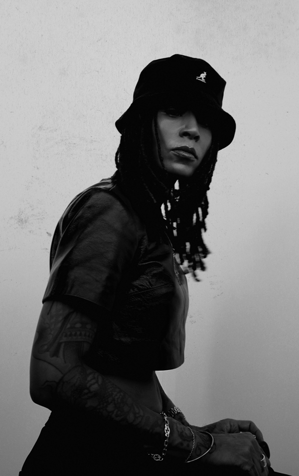 A woman wearing a black bucket hat, black leather jacket, and showing tattoos on her arm, posed against a plain background in a black-and-white photo.