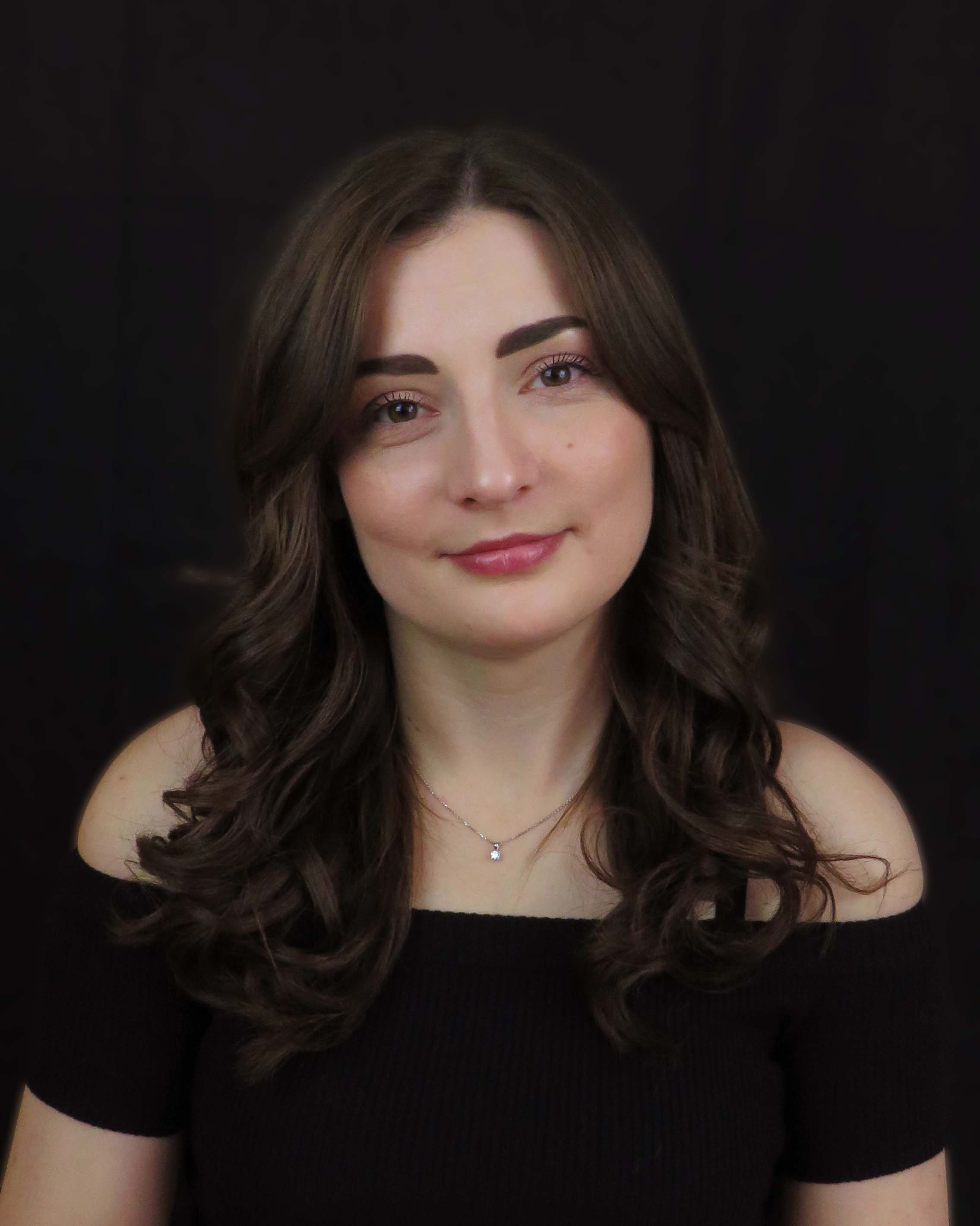 A young woman with long, wavy brown hair wearing a black off-shoulder top and a delicate necklace against a dark background.