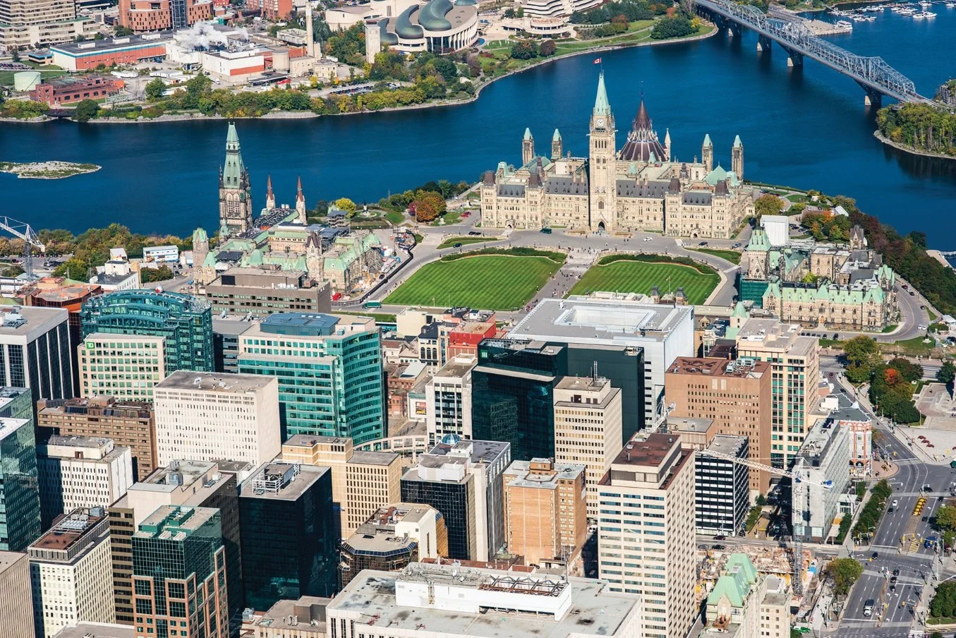 Aerial view of Ottawa, Canada, showcasing Parliament Hill with its historic buildings and green lawns, situated beside the Ottawa River with bridges and city infrastructure surrounding it.