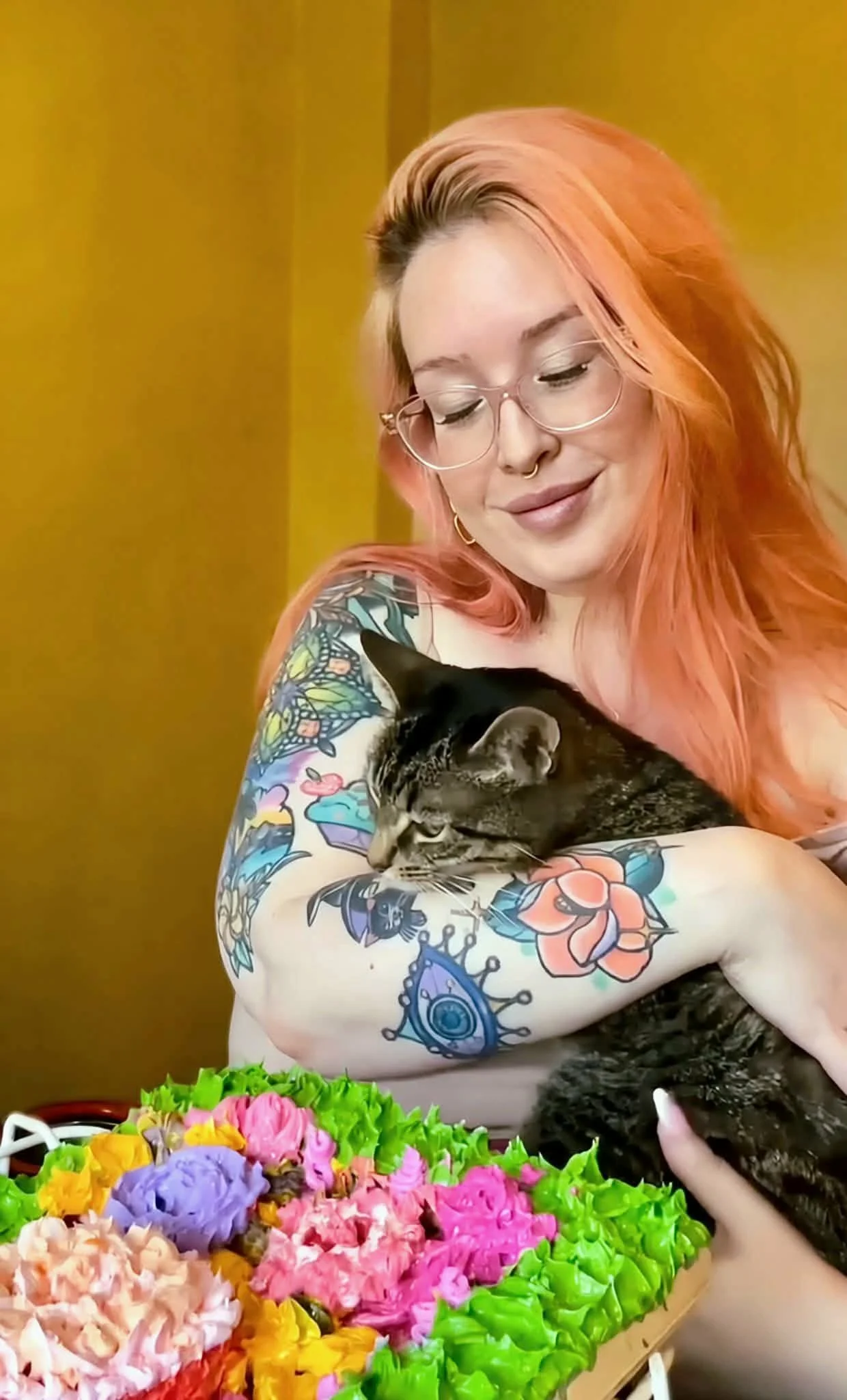 A woman with pink hair, glasses, and colorful tattoos on her arm is holding a black and brown tabby cat. There are vibrant flowers in the foreground and a yellow wall in the background.