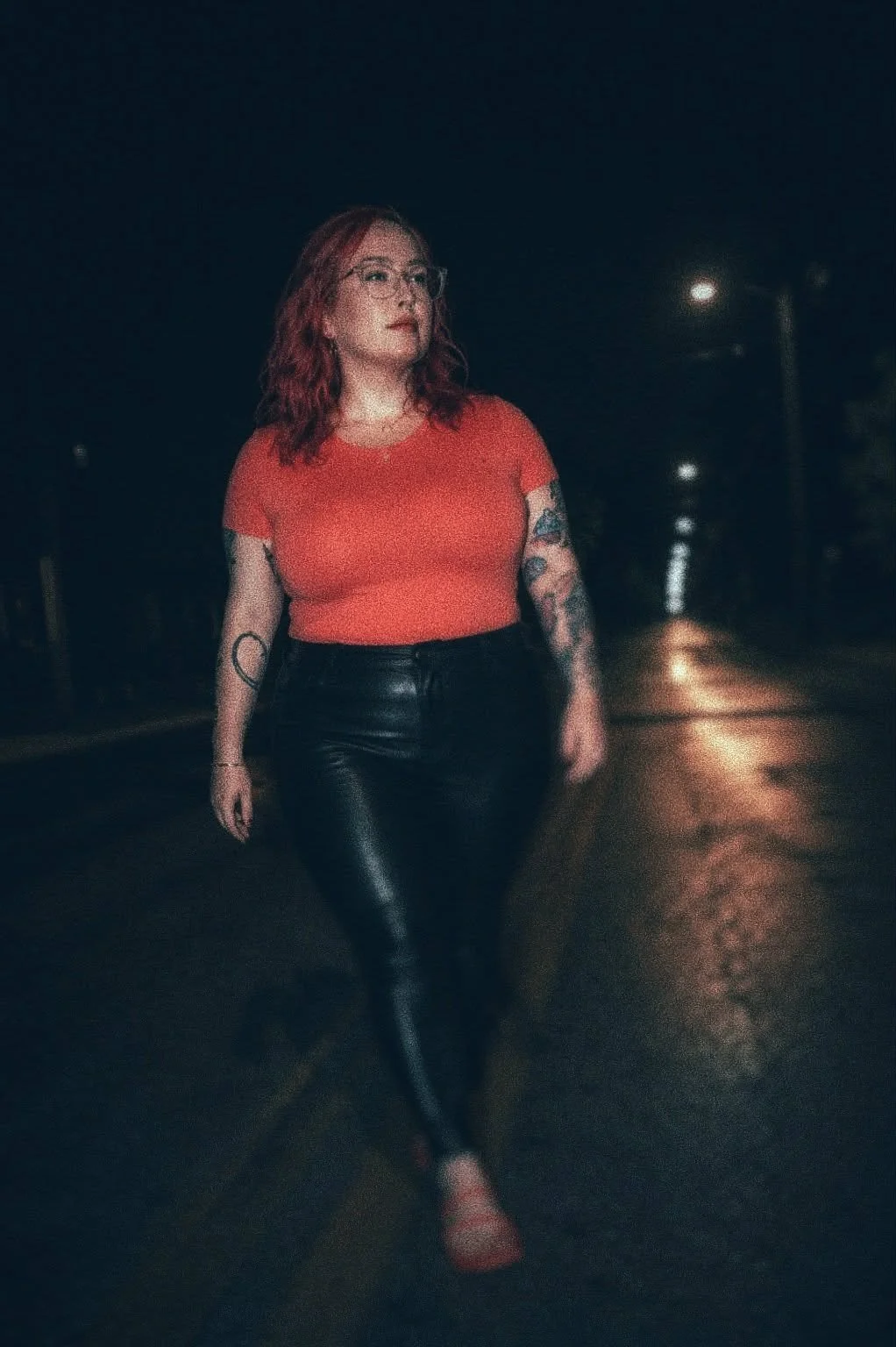A woman with red, wavy hair and glasses walks on a dark, wet street at night. She is wearing a red top, black leather pants, and red shoes, and has tattoos on her arms.