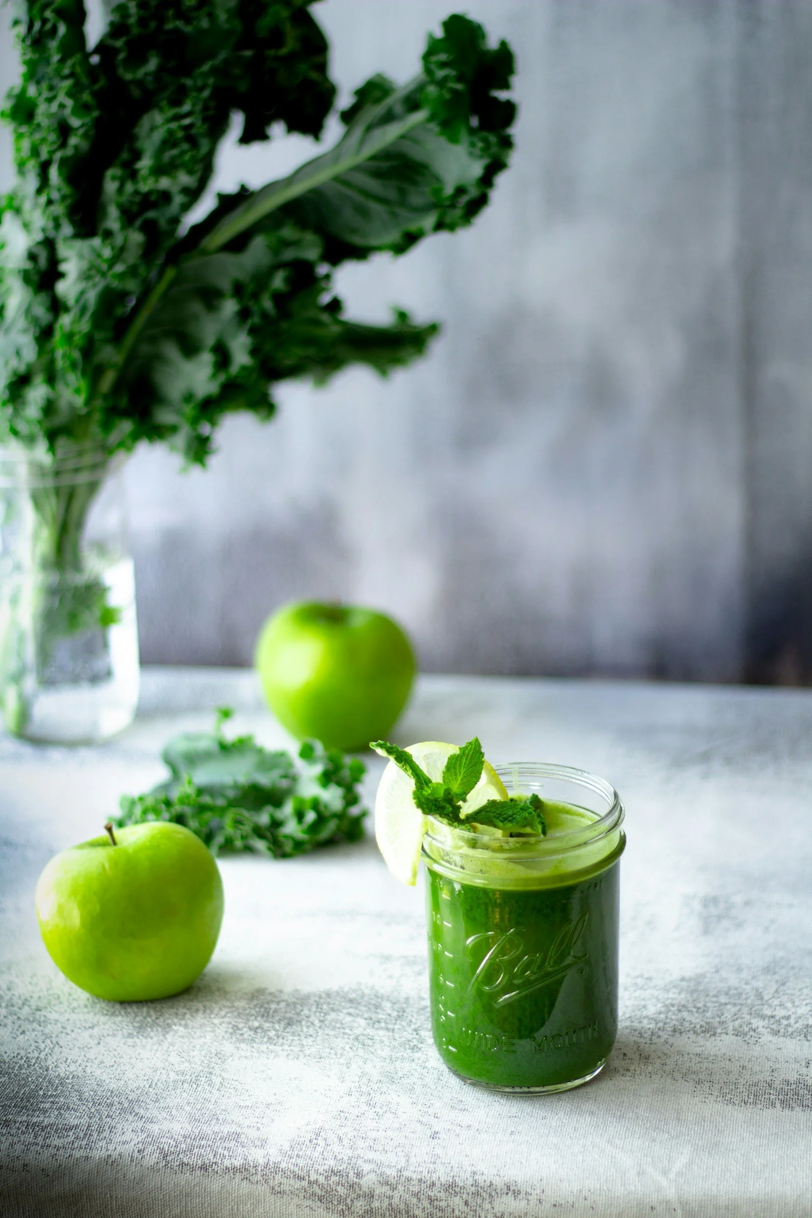 A green smoothie in a Mason jar with mint and lemon slices, placed on a white table with green apples and leafy greens in the background.