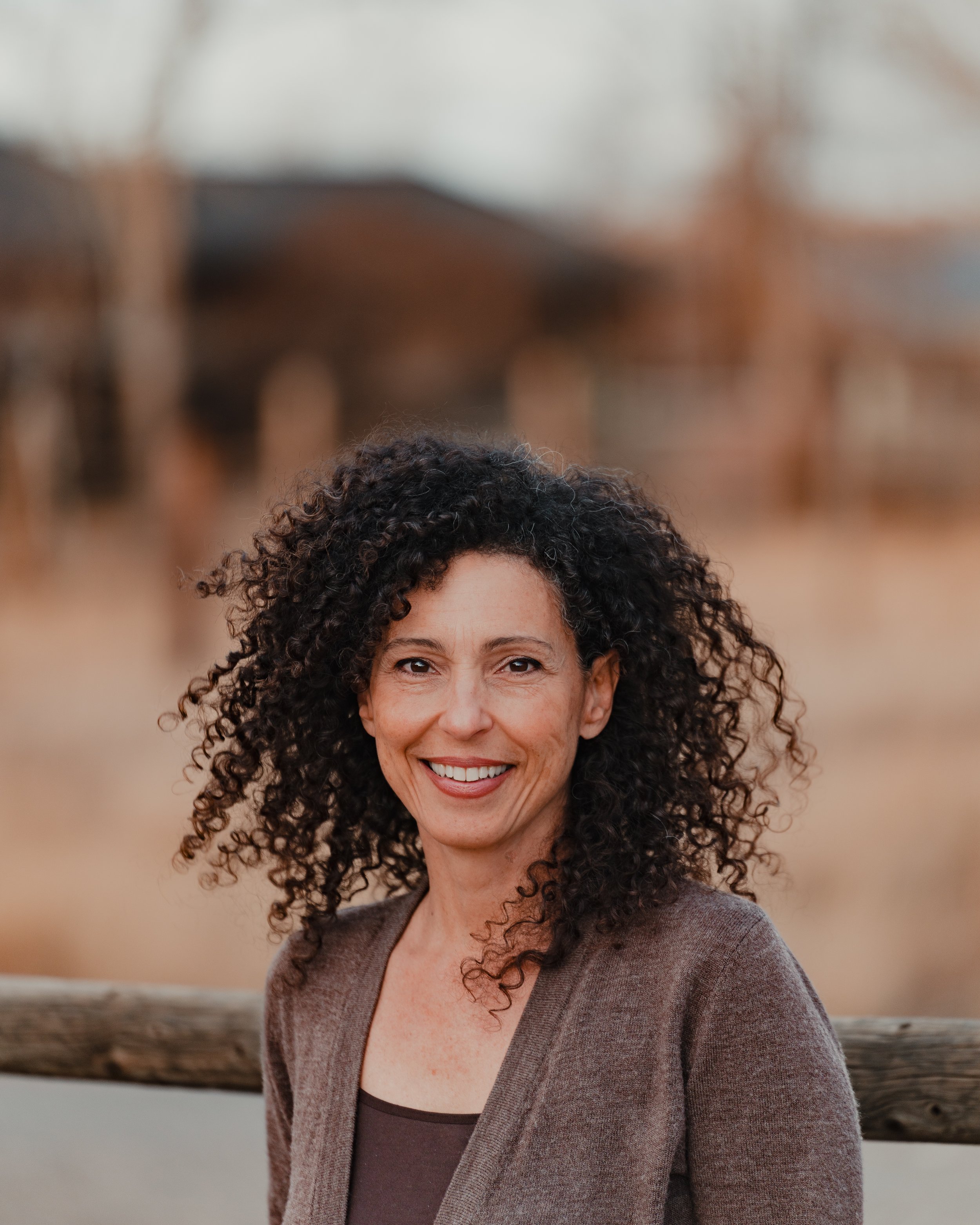 A woman with curly dark hair smiling outdoors with a wooden fence and blurred background.