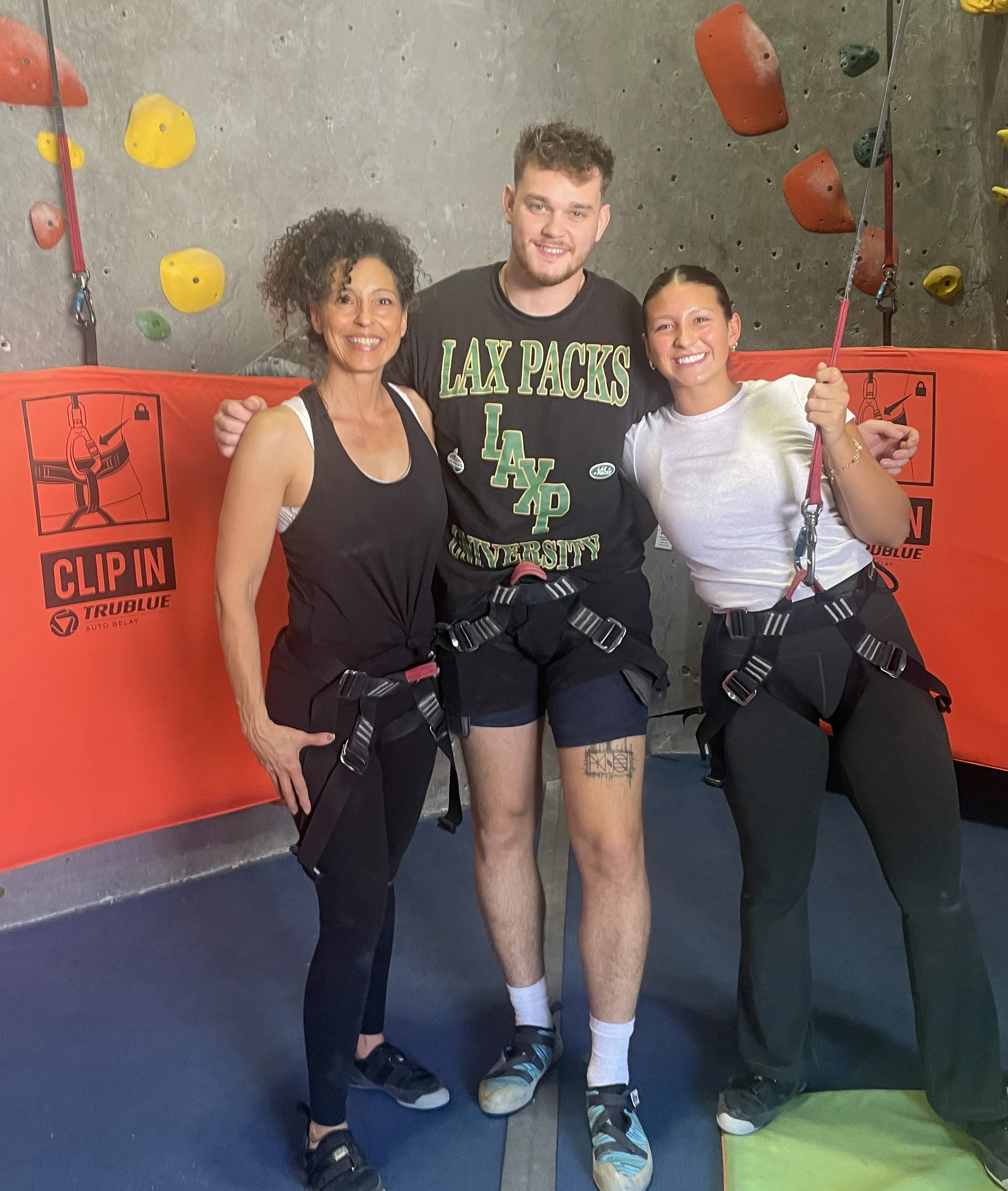 Three people wearing harnesses and climbing shoes standing in front of an indoor climbing wall with colorful holds. They are smiling, with the person in the middle having their arms around the other two.
