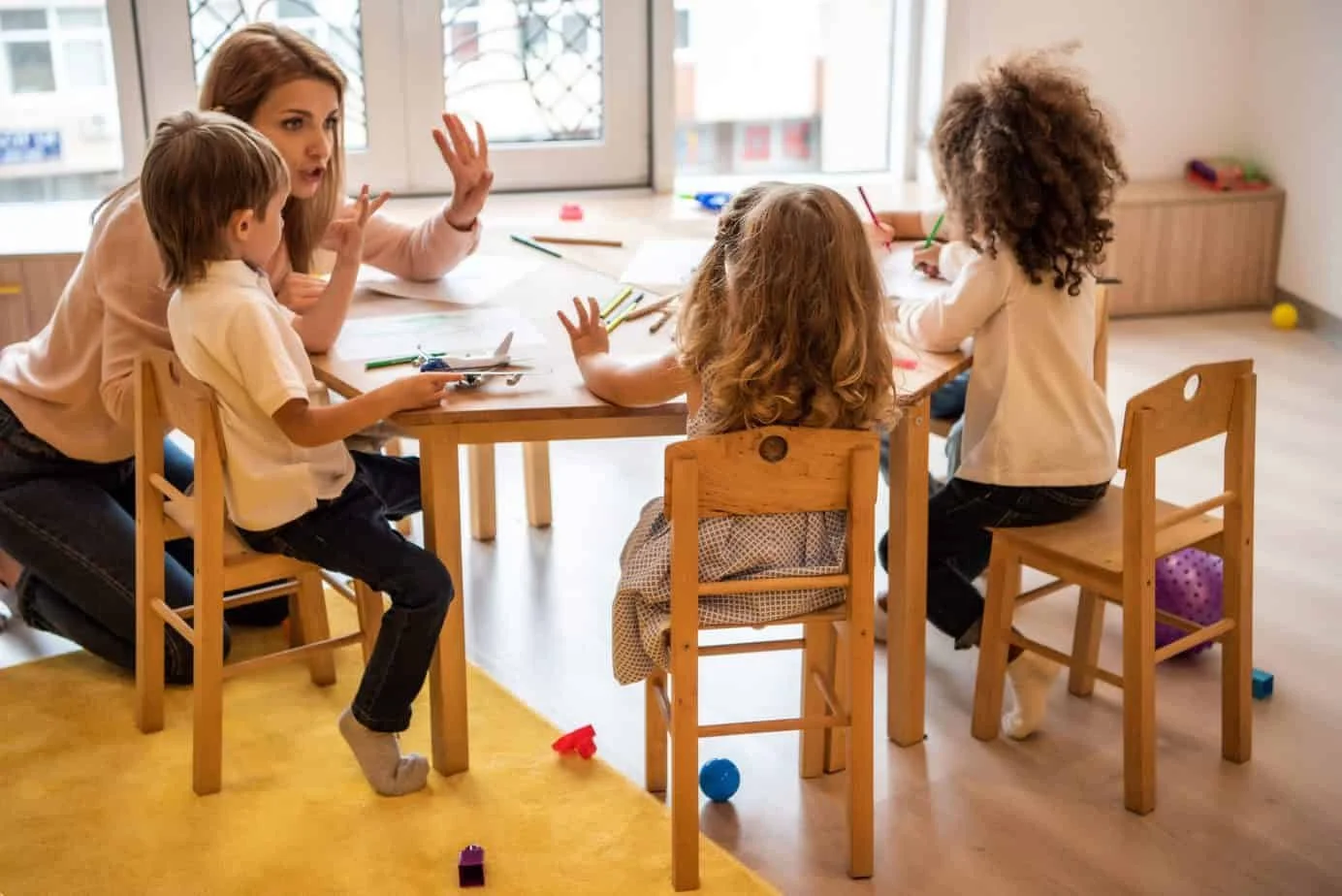 A teacher with four young children sitting around a table in a classroom, engaging in a discussion or lesson. The children have toys and art supplies in front of them.