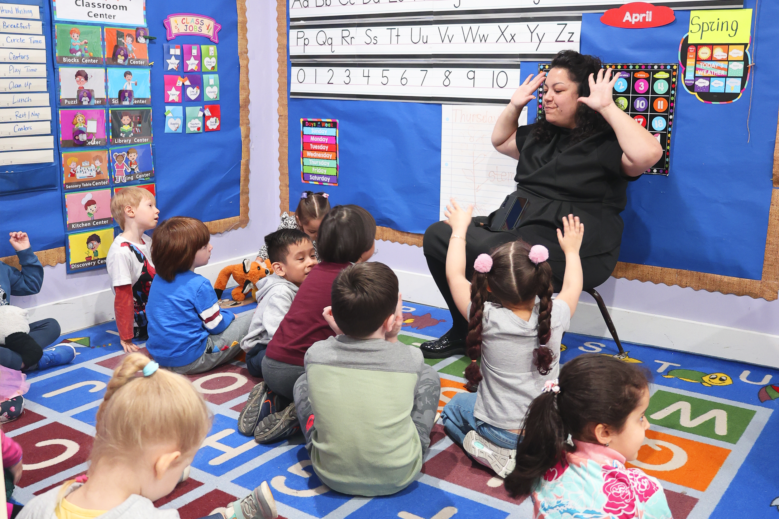A teacher is sitting on a stool at the front of a preschool classroom. She is engaging with young children sitting on a colorful alphabet rug, some with hands raised. The classroom has blue bulletin boards with educational charts, seasonal and activity displays, and a whiteboard with handwriting practice.