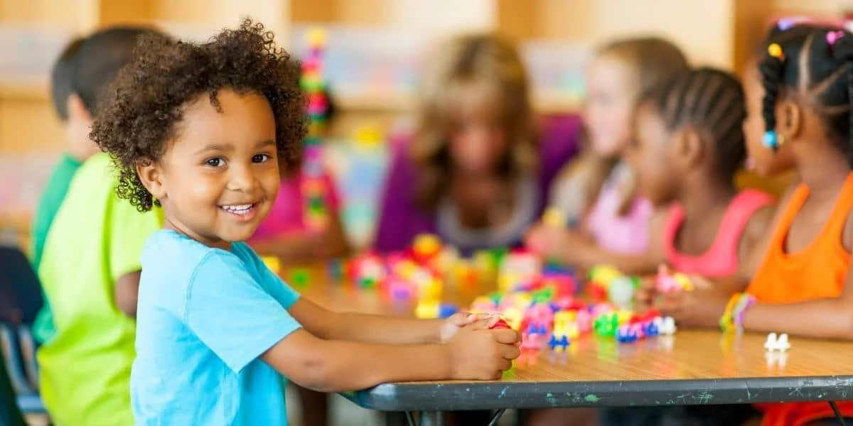 A young girl with curly hair smiling at the camera while sitting at a table with other children, playing with colorful building blocks in a classroom setting.