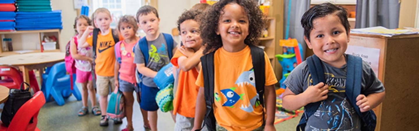 A group of children standing in line inside a classroom, smiling and carrying backpacks.