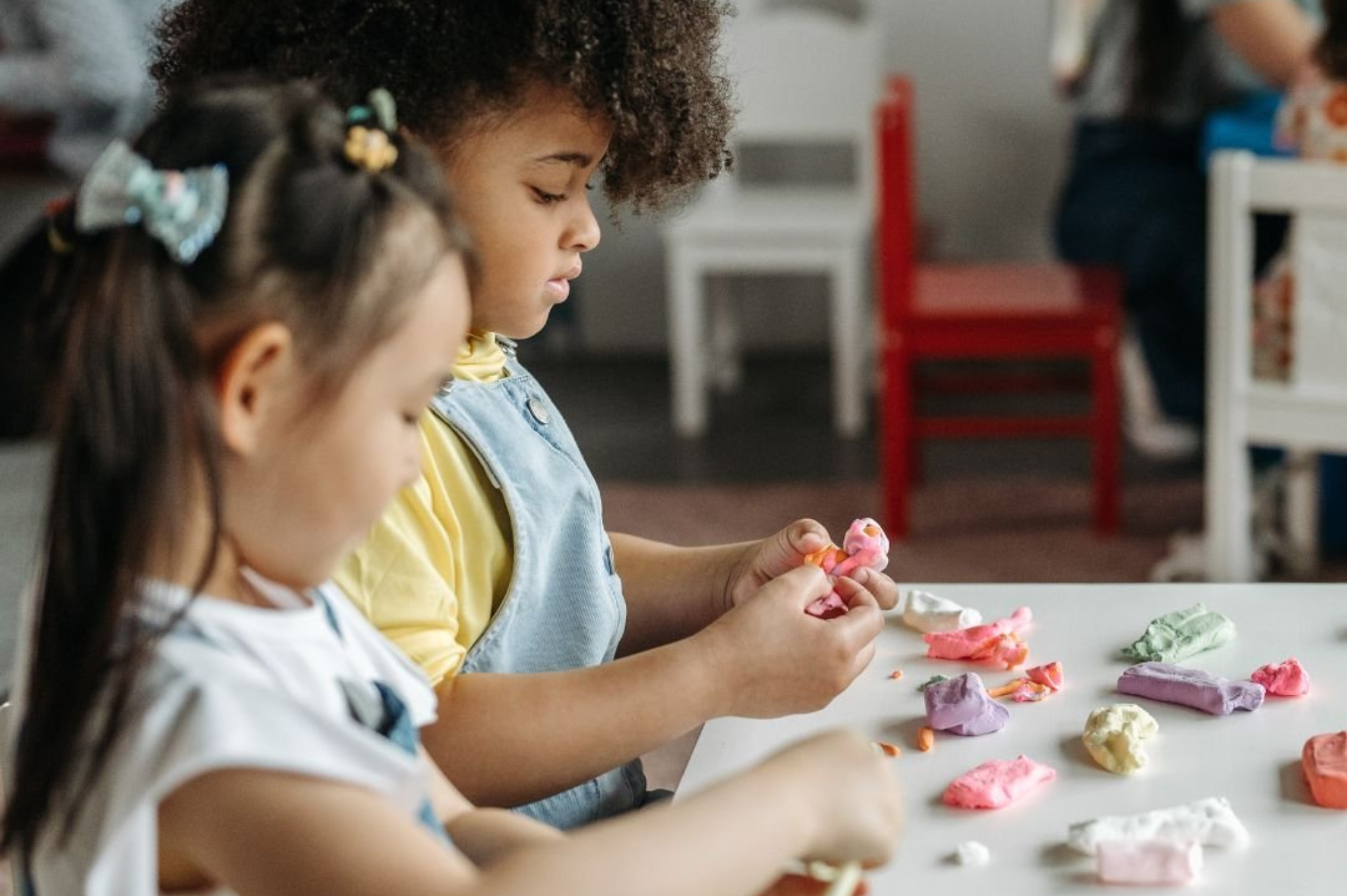 Two young girls playing with colorful playdough at a white table in a classroom.