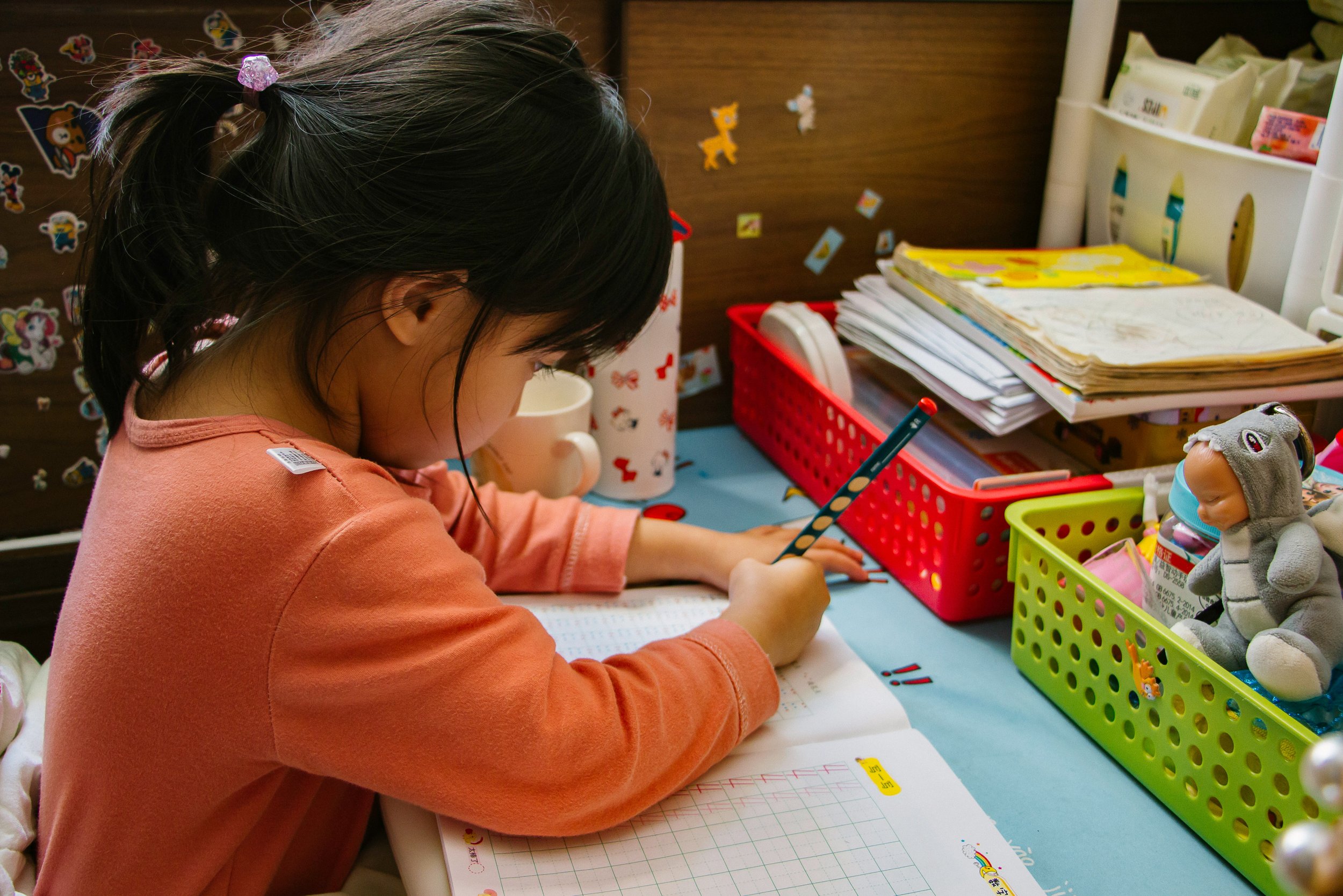 A young girl with dark hair in a ponytail, wearing an orange shirt, sitting at a desk and writing in a notebook with a pen. The desk has organized supplies in colorful baskets, including a stuffed animal, papers, and stationery, with a decorated background.