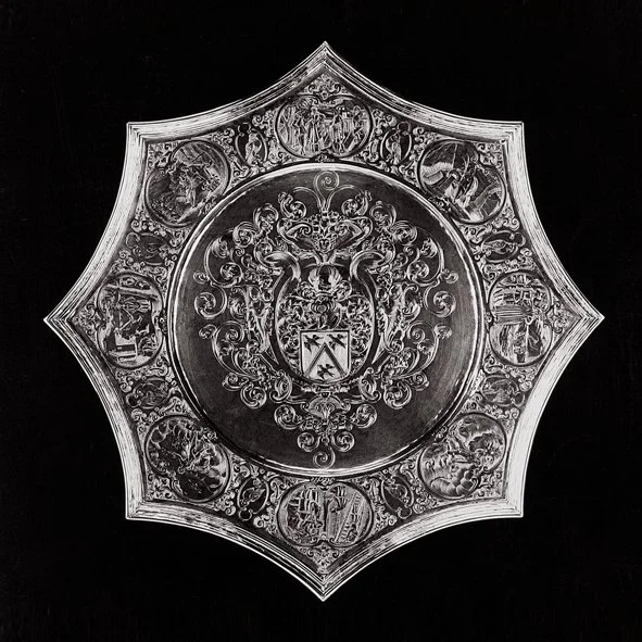 Decorative silver shield with intricate engravings and crest in the center, surrounded by smaller engraved medallions, set against a black background.