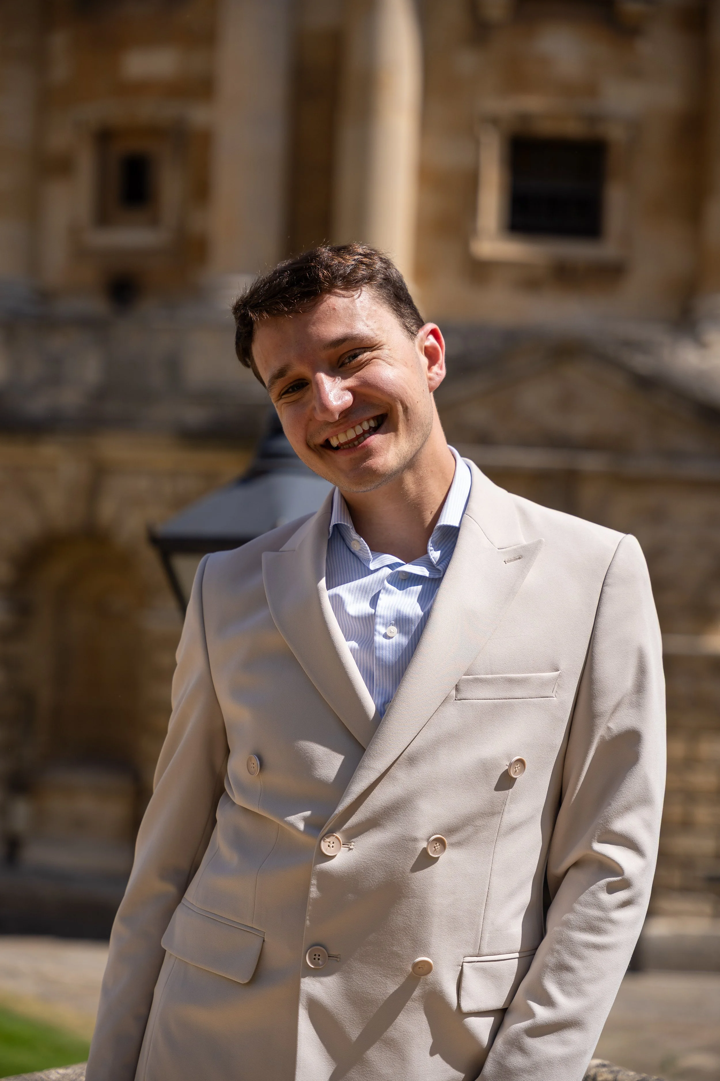 Young man smiling in beige double-breasted suit in front of historic building with stone facade and windows.