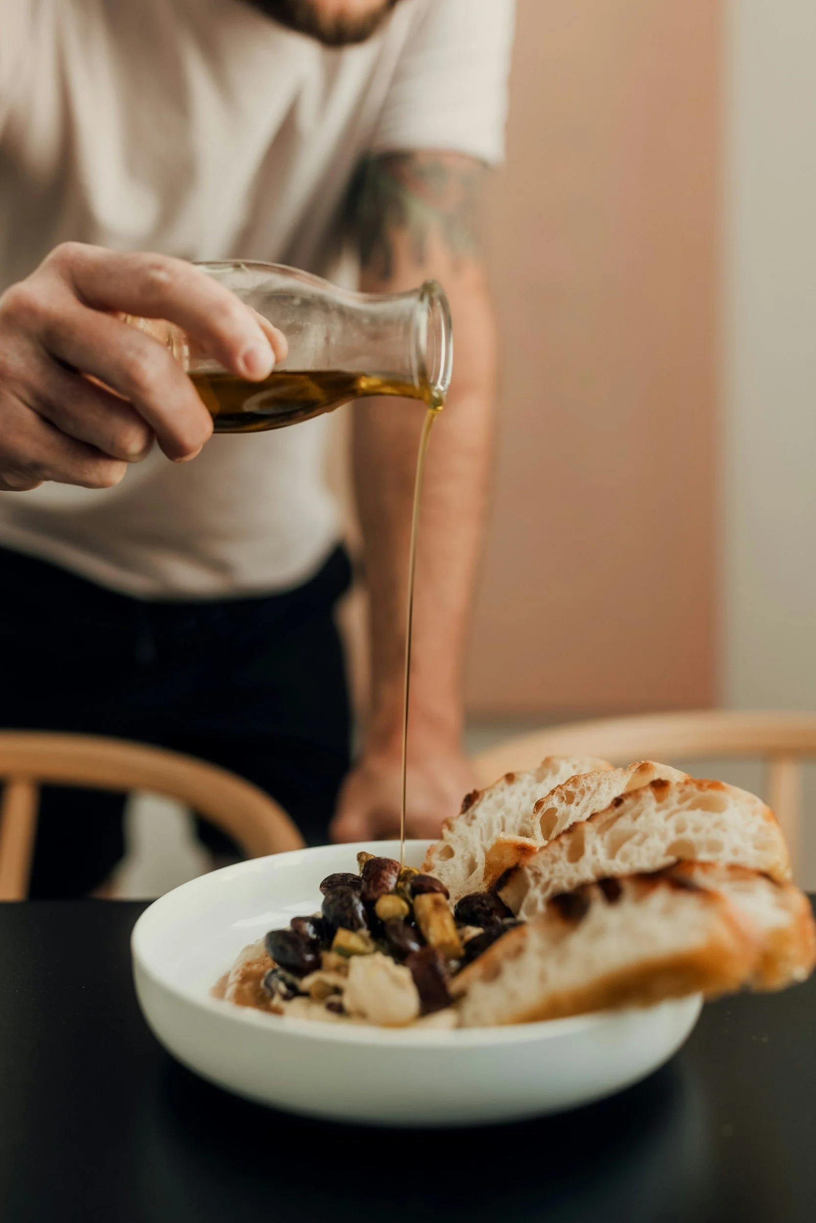 Una persona vierte aceite de oliva sobre un plato de hummus con pan árabe y una mezcla de verduras picadas y frijoles.