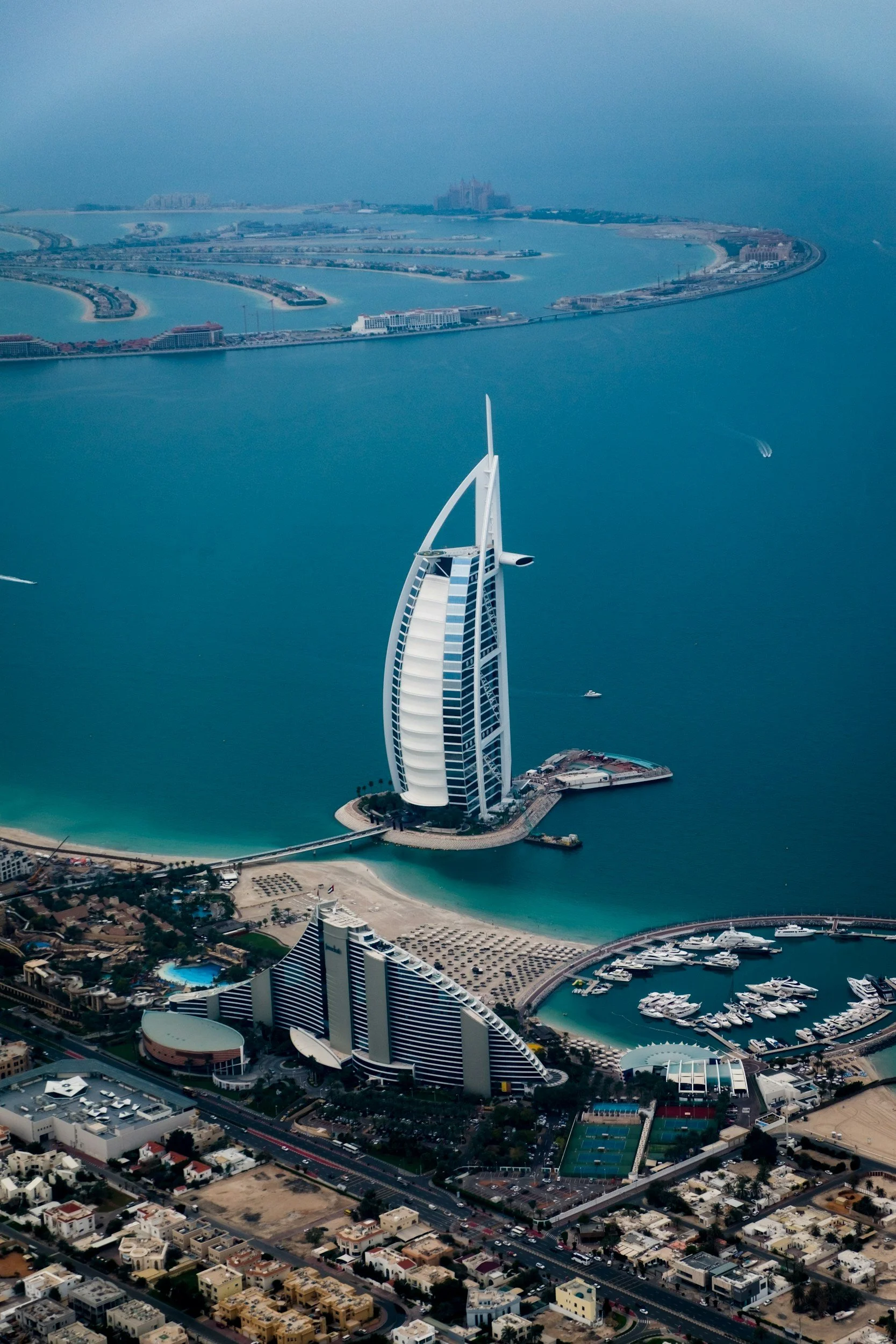 Vista aérea del hotel Burj Al Arab en Dubai, situado en una isla artificial en el mar con barcos cerca, con la ciudad y la playa al fondo.