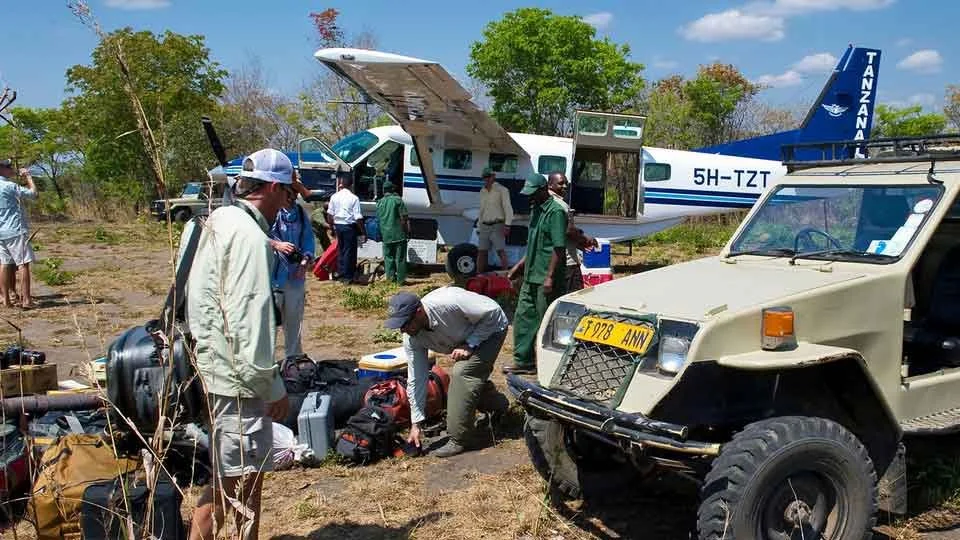 Fly fishing safari guests and staff unloading gear from a bush plane next to a safari vehicle in Tanzania, ready for tigerfish adventure.