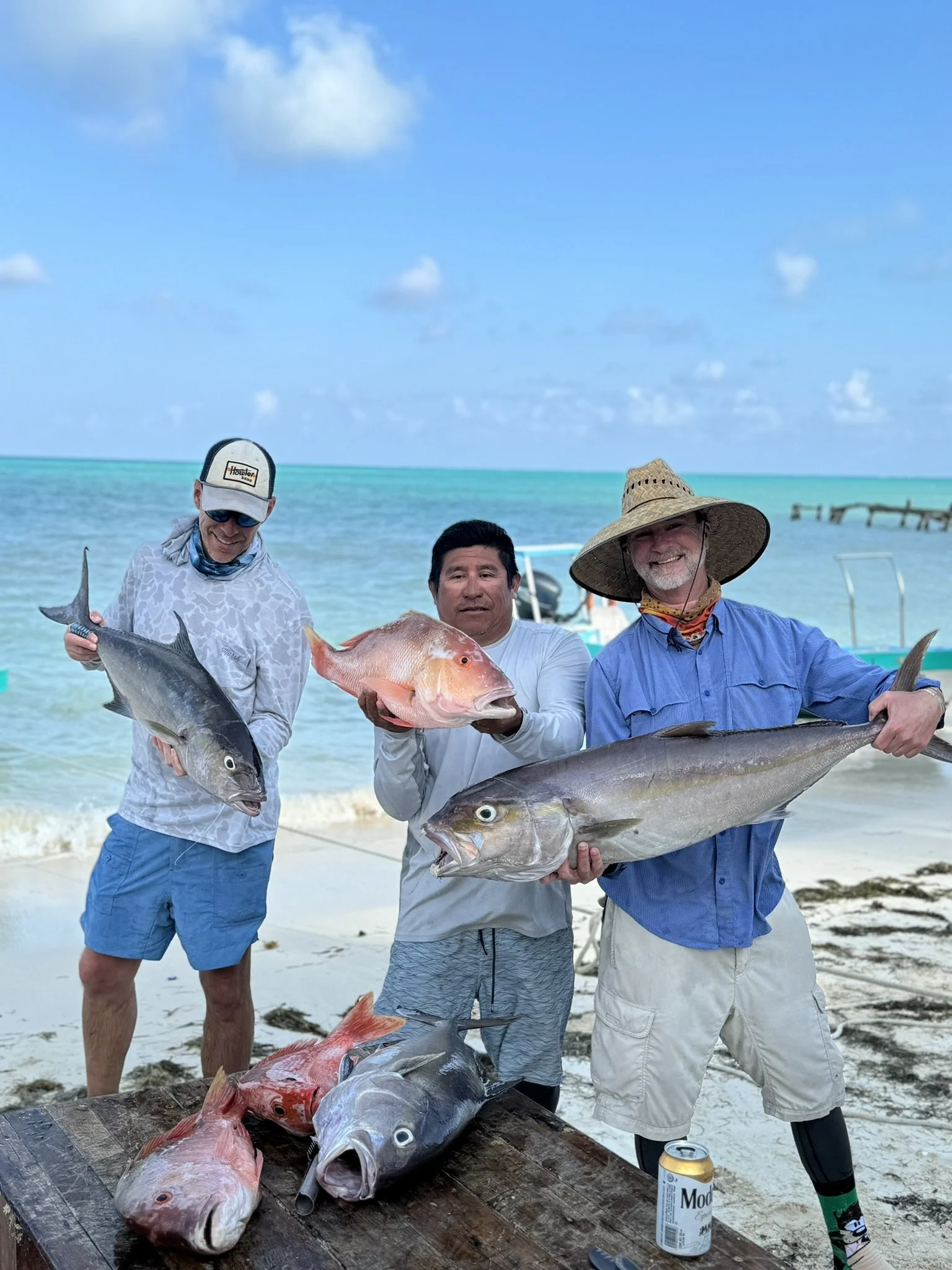 Three men standing on a beach holding large fish, with a table of more fish in front and boats in the background at the ocean.