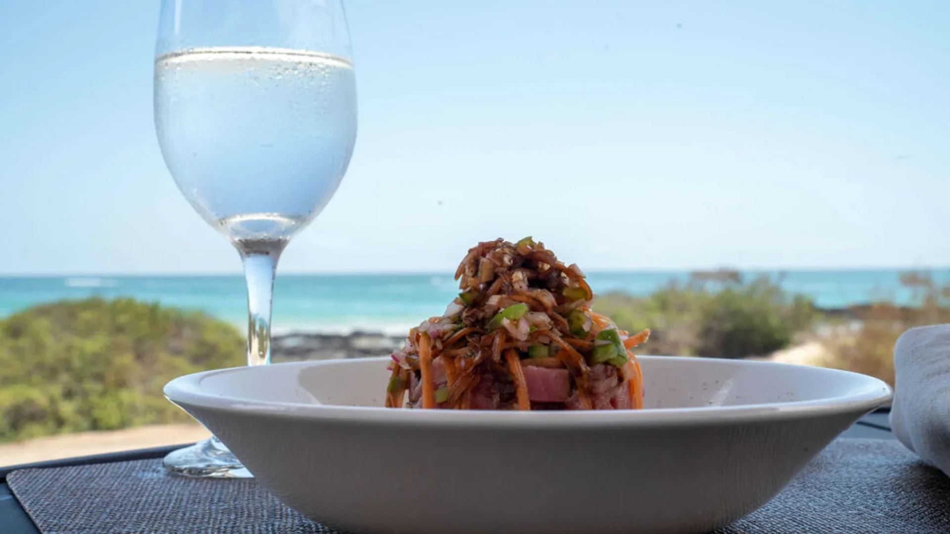A plate of salad with shredded vegetables near the beach, with a glass of water or clear beverage and ocean in the background.