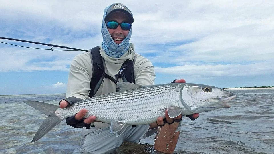 Angler wearing a hat and face covering, kneeling in the shallow waters of Farquhar Atoll, Seychelles, holding a bonefish.
