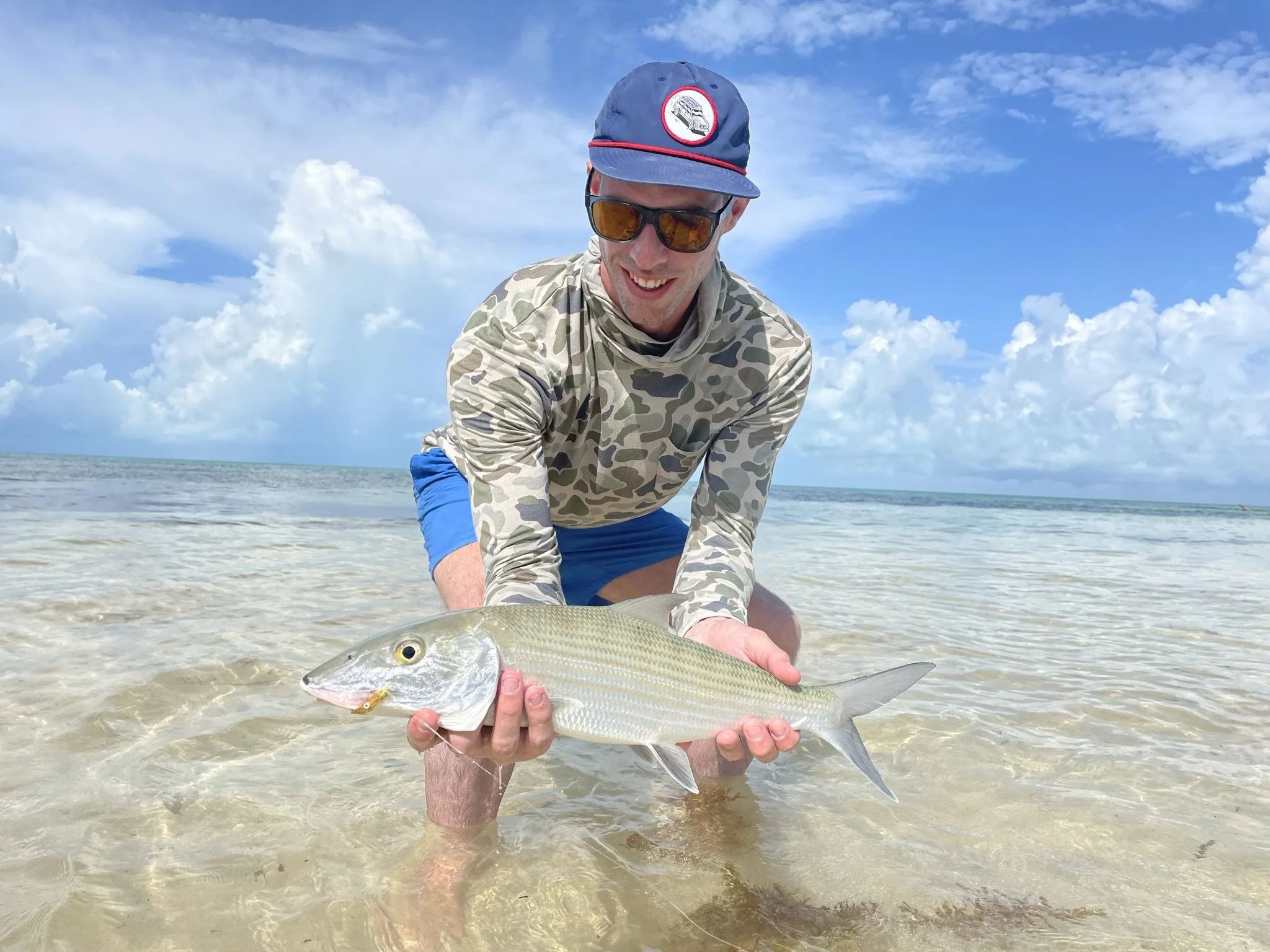 Man in camouflage shirt, blue shorts, wearing sunglasses and a cap, kneeling in shallow waters of Ascension Bay, Mexico holding a bonefish.