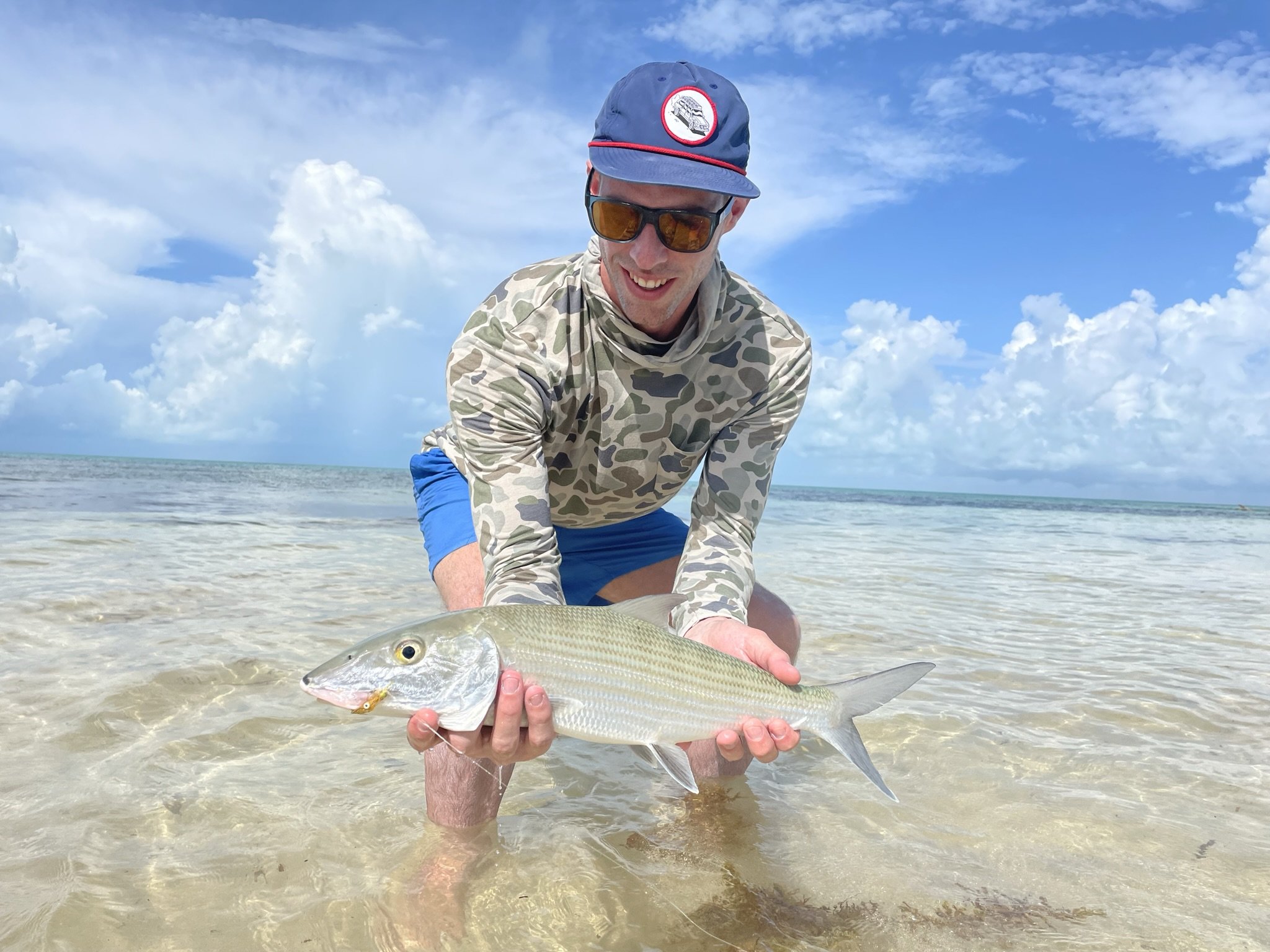Man in camouflage shirt, blue shorts, wearing sunglasses and a cap, kneeling in shallow ocean water holding a fish he caught.