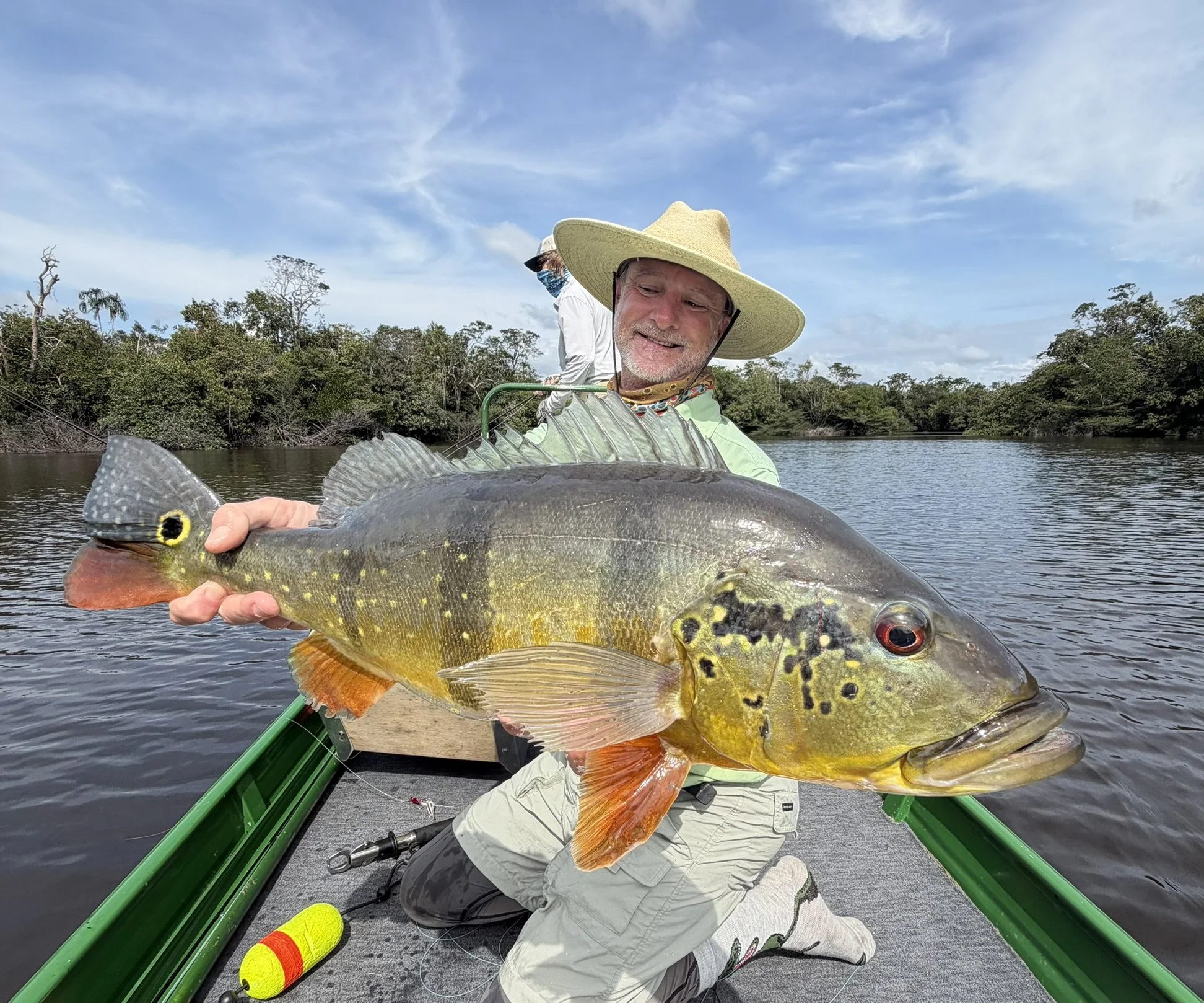 Angler wearing a wide-brim hat holding a large peacock bass on the Agua Boa River surrounded by tropical trees in Brazil