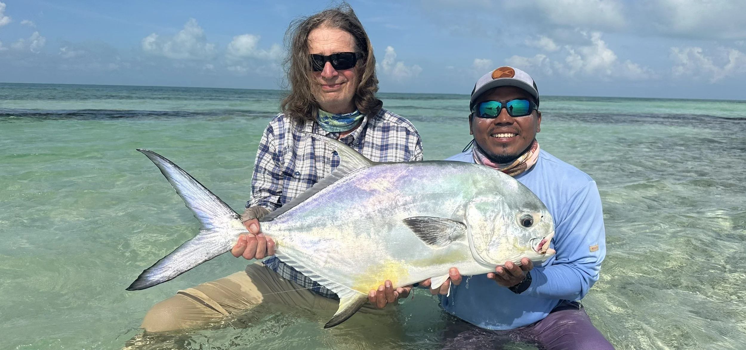 Two men in water at the beach holding a large fish, smiling, with blue sky and ocean in the background.