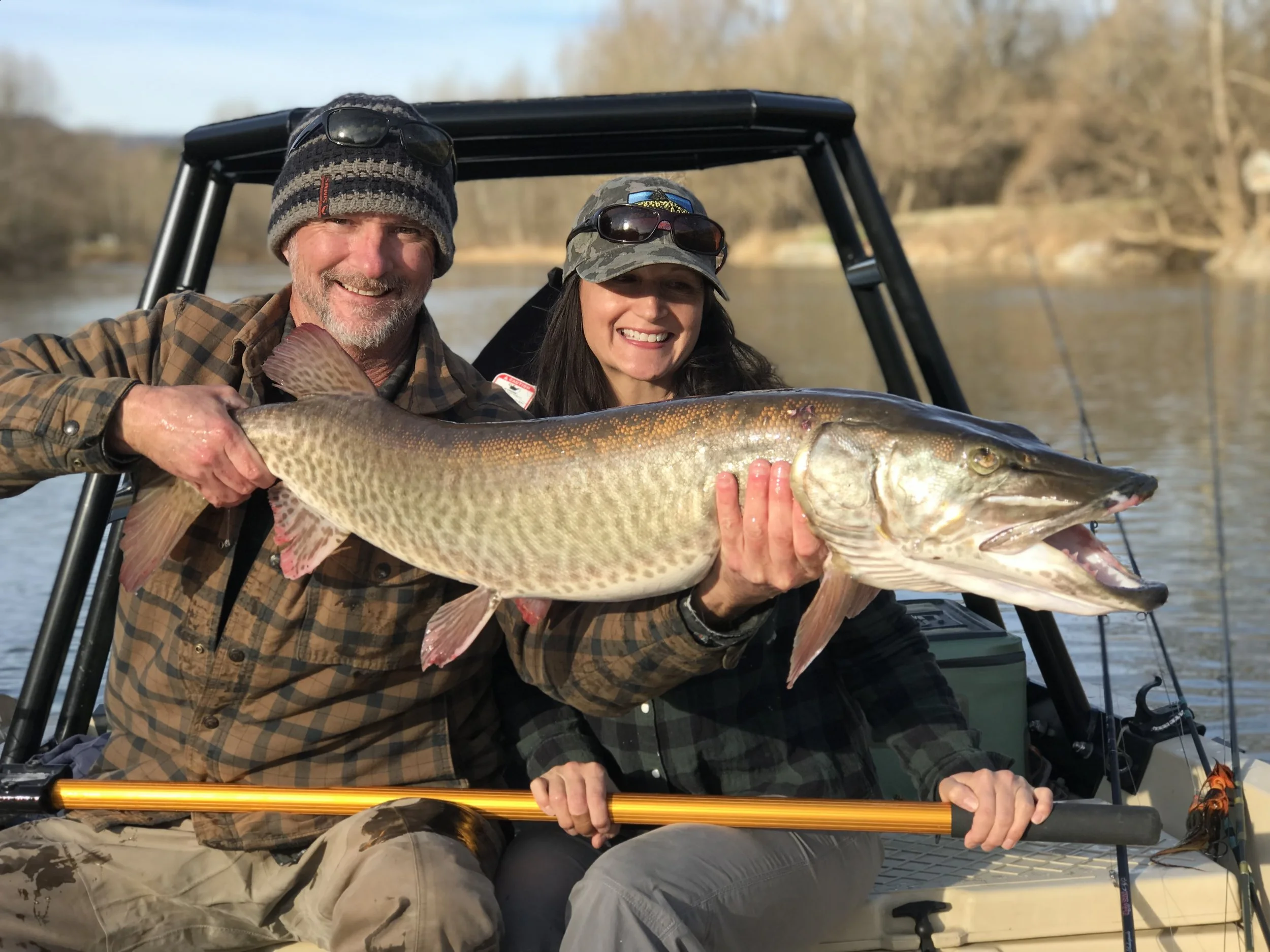 A man and woman on a boat holding a large fish, likely a pike, with the man smiling and the woman smiling behind it.