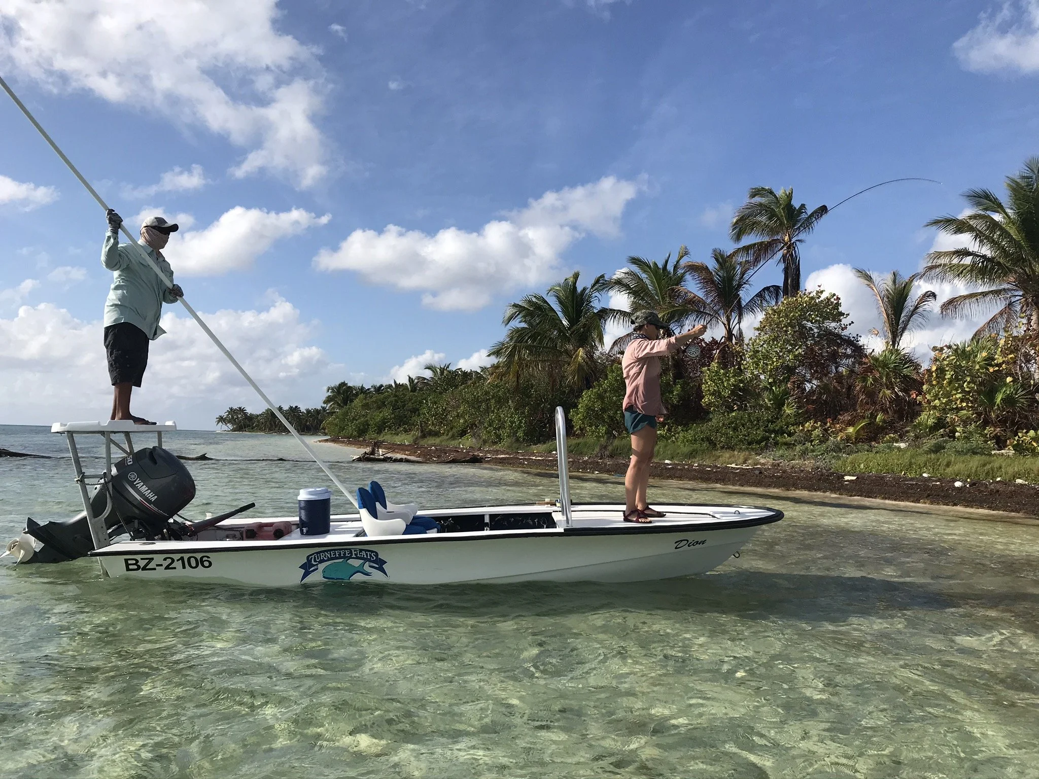 Angler standing on the bow of a Turneffe Flats skiff fighting a fish while the guide stands on the poling platform, with tropical mangroves in the background.