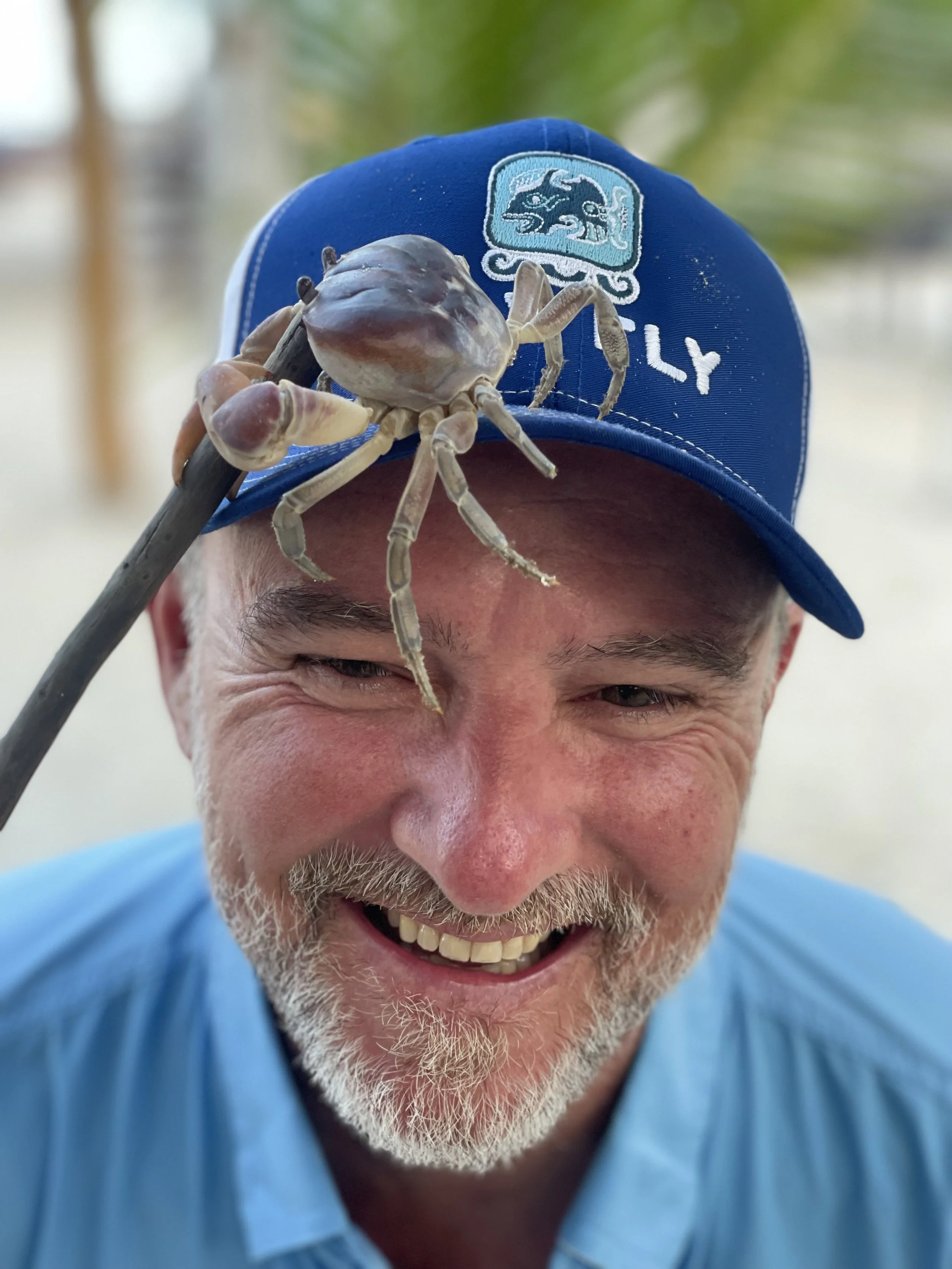 A man smiling while posing with a crab on his hat, outdoors with blurred greenery and a sandy background.