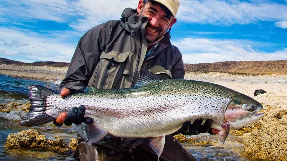 Angler holding a massive rainbow trout on the shoreline of Jurassic Lake in Argentina during a fishing trip at Estancia Laguna Verde Lodge.