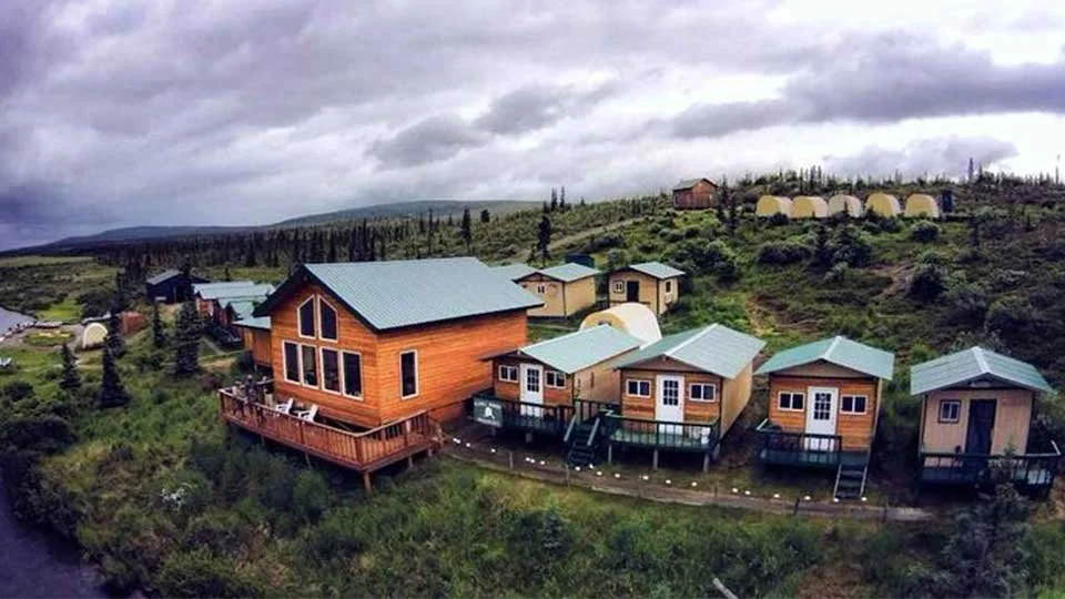 A scenic view of a hillside with small wooden houses, some with decks, under a cloudy sky, surrounded by greenery and trees.