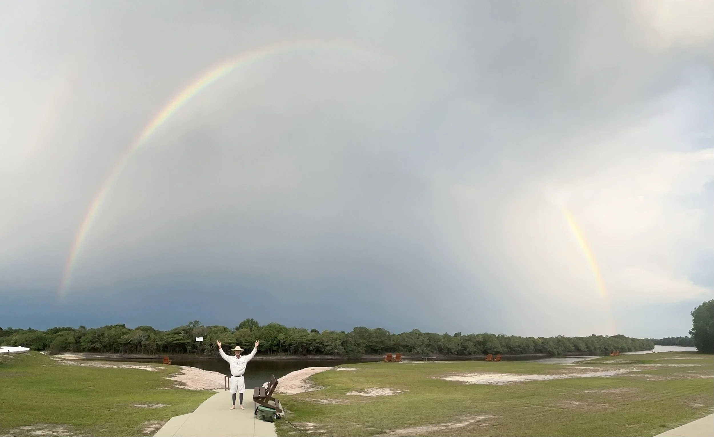Person standing with arms raised under a double rainbow in a park with grass and trees.